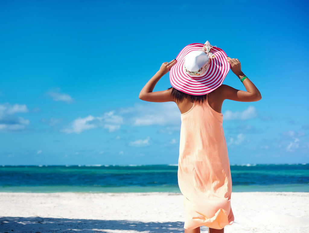Hot beautiful woman in colorful sunhat and dress walking near beach ocean on hot summer day on white sand